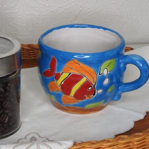 May include: A blue ceramic coffee mug with a colorful fish design. The fish has orange, red, and yellow scales. The mug has a white interior and a handle.