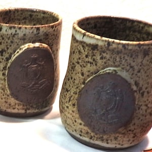 May include: Two brown and white ceramic mugs with a brown circular design on the front. The mugs have a speckled glaze.