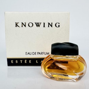 May include: A miniature perfume bottle with a black lid, containing a golden liquid. The bottle is in front of a white box with the word "KNOWING" printed on it. The box also has the words "EAU DE PARFUM" and "ESTEE LA" printed on it.