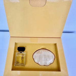 Peut inclure: Un coffret cadeau de parfum dans une boîte dorée. L'ensemble comprend un petit flacon de parfum en verre avec un bouchon noir et un savon blanc en forme de coquillage. Le flacon de parfum porte du texte.