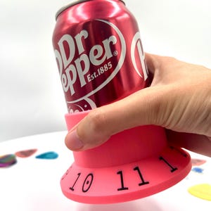 May include: Pink guitar pick holder with numbers 10 and 11 visible. A can of Dr Pepper is being held in the holder.