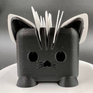 May include: Black cat-shaped utensil holder with white ears and a white bristle brush for cleaning. The holder has two round holes for eyes and a small nose.