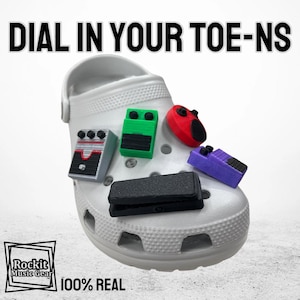 May include: A white shoe with five colorful guitar pedal-shaped charms attached to it. The charms are green, black, red, purple, and black. The shoe has the text "DIAL IN YOUR TOE-NS" above it and "Rockit Music Gear" and "100% REAL" below it.