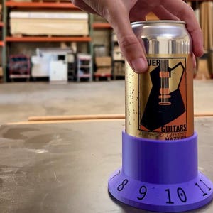 Könnte beinhalten: Ein lilafarbener, 3D-gedruckter Dosenhalter mit den Zahlen 8, 9, 10 und 11 um die Basis. Eine Bierdose mit einem gold-schwarzen Etikett sitzt im Halter. Das Etikett lautet "Guitars Hybrid Theory Hazy IPA".