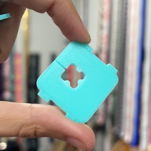 May include: A square, light blue plastic object with a cross-shaped hole in the center. The object has a textured surface and small tabs on each corner. The object is held by a person's fingers.