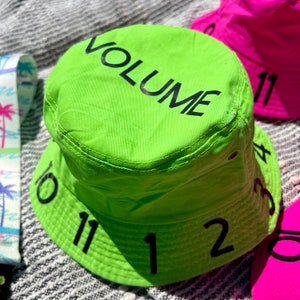 May include: A bright green bucket hat with the word "VOLUME" printed on the crown. The brim of the hat has numbers 10, 11, 1, 11, 12, and 3 printed on it.