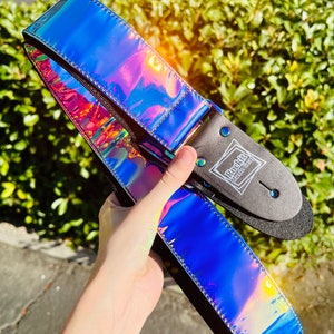 May include: A guitar strap with a vibrant, iridescent blue finish. The strap has a black backing and is stitched with a silver thread. The strap has the text "Rock It Music Gear" printed on it.