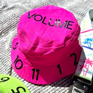 May include: A pink bucket hat with the word "VOLUME" printed on the crown and numbers 9, 10, 11, 12, and 2 printed around the brim.