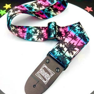 May include: A guitar strap with a vibrant tropical design. The strap features a repeating pattern of black palm trees against a gradient of pink, blue, and white. The strap has a black adjustable buckle and a black leather end piece with the "Rockit Music Gear" logo.