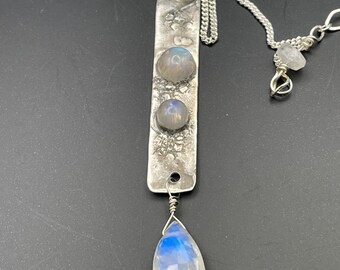 May include: A silver necklace with a long, rectangular pendant featuring three round moonstone gems and a teardrop-shaped moonstone pendant.