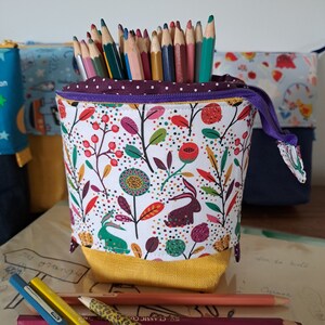 Peut inclure: Un porte-crayons en tissu blanc avec un motif floral coloré et un fond jaune. Le porte-crayons est rempli de crayons de couleur et a une fermeture éclair violette.