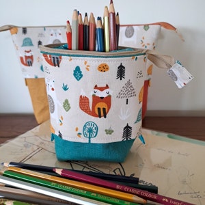 Peut inclure: Un porte-crayons en turquoise et beige avec une fermeture éclair. Le porte-crayons est décoré d'un motif répété de renards, d'arbres et de feuilles. Le porte-crayons est rempli de crayons de couleur.