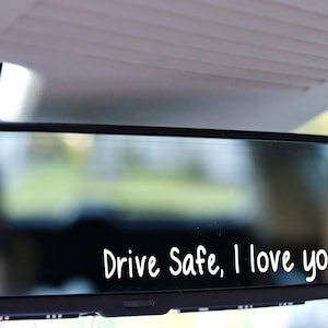 May include: A rearview mirror in a car with the words "Drive Safe, I love you" written on the glass.