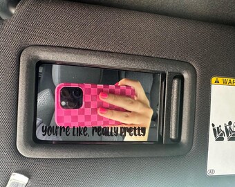 May include: A pink checkered phone case is reflected in a car visor mirror. The mirror has the text "you're like, really pretty" in a black script font. The visor is black and has a warning label.