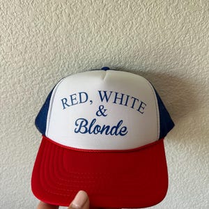 May include: A red, white, and blue trucker hat with the text "RED, WHITE & BLONDE" in blue script. The hat features a red brim, a white front panel, and a blue mesh back.