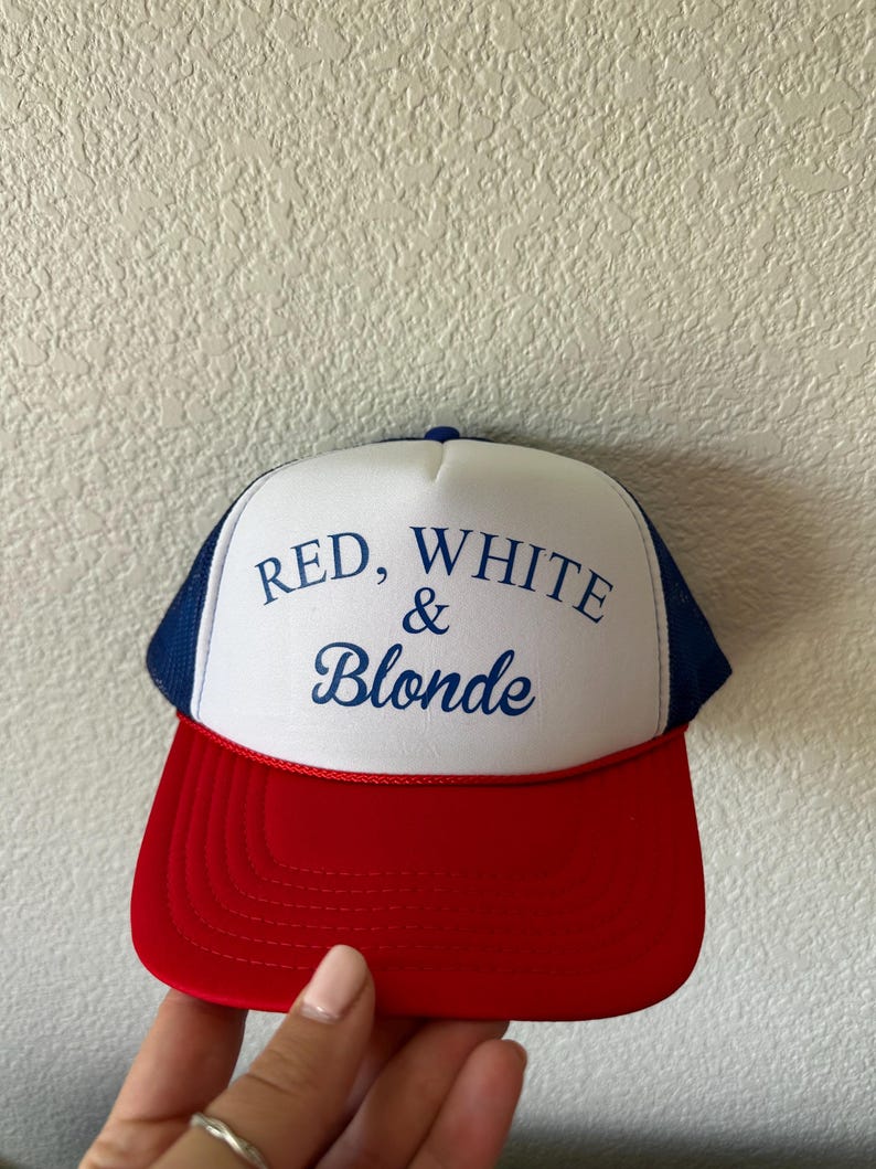 May include: A red, white, and blue trucker hat with the text "RED, WHITE & BLONDE" in blue script. The hat features a red brim, a white front panel, and a blue mesh back.