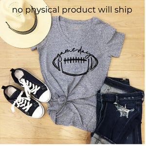 May include: A heather gray V-neck t-shirt with a black graphic of a football and the text "game day" in cursive script, paired with a straw hat, black canvas sneakers with white laces, and dark blue distressed denim jeans.