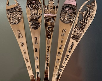 May include: A collection of silver souvenir spoons, each engraved with a different state name: Arizona, Idaho, Chicago, Minnesota, and Des Moines. The spoons feature detailed designs of landmarks and state symbols.