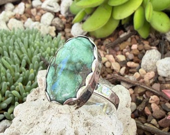 May include: A silver ring featuring a large, oval-shaped green stone with dark inclusions. The stone is set in a scalloped silver bezel, and the band is made of polished silver. The ring is displayed on a textured, light-colored rock surface.