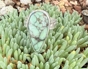 May include: A teardrop-shaped ring featuring a pale mint green stone with prominent dark brown veining. The stone is set in a polished silver-toned metal band with a hammered texture along the sides, showcasing a distinct organic pattern.