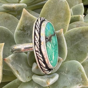 May include: A silver ring featuring an oval, turquoise-colored stone with dark veining. The stone is set in a decorative bezel with a leaf-like pattern. The ring is displayed against a backdrop of green succulent leaves.