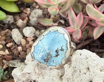 May include: A silver ring featuring a large, irregular, light blue stone with dark brown matrix inclusions. The stone is set in a scalloped silver bezel, showcasing a smooth, polished surface and a distinct, organic shape that highlights the natural variations of the gemstone.