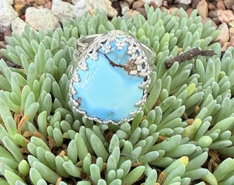 May include: A silver ring featuring a large, pear-shaped light blue turquoise stone with a brown matrix inclusion. The stone is set within a decorative, scalloped silver bezel with intricate metalwork detailing along the outer edge of the setting.