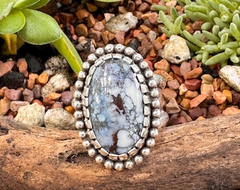 May include: Oval-shaped silver ring with a large, marbled stone. The stone features shades of blue, white, and brown, set in a silver bezel. The ring has a beaded border and is displayed on a piece of wood.