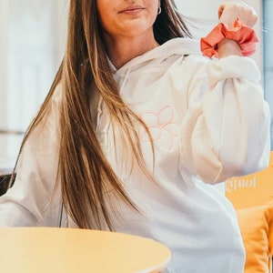 Op de afbeelding: Een jonge persoon draagt een witte hoodie met een roze haarband. Ze zitten aan een gele tafel in een café.