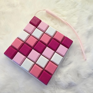 May include: A square-shaped grid of twenty-five individual square keys arranged in a five-by-five pattern. The keys feature a textured surface in varying shades of pink, magenta, and white. A thin, light pink cord is attached to the top corner.