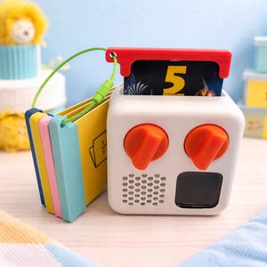 May include: A white cube-shaped toy with orange knobs and a red handle, showing a card with the number 5. A small stack of colourful cards is attached with a green cord. The toy has a speaker and a black square.