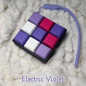 May include: A black tray containing nine square blocks in shades of purple, white, and pink. A matching purple strap is attached. The text "Electric Violet" is visible below the tray.