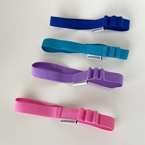 Könnte beinhalten: Vier farbige elastische Stirnbänder in Blau, Türkis, Lila und Pink. Jedes Stirnband hat ein dekoratives, plissiertes Design und ein kleines weißes Etikett mit Text. Die Stirnbänder sind auf einer weißen Oberfläche angeordnet.