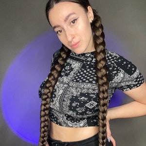 May include: A woman with long brown braids wearing a black and white bandana print crop top and black jeans.