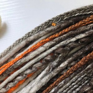 Peut inclure: Un ensemble de dreadlocks synth&eacute;tiques dans diff&eacute;rentes nuances de gris, de brun et d'orange. Les dreadlocks sont tress&eacute;es et ont une apparence textur&eacute;e.