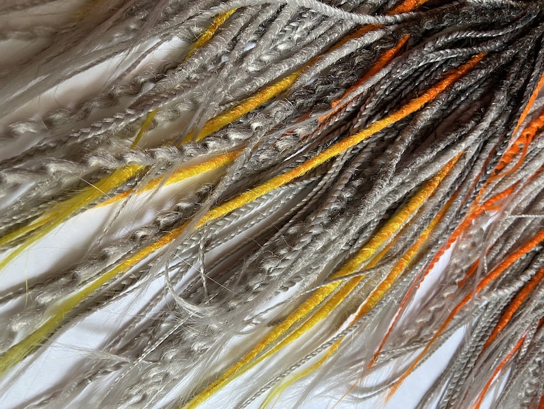 Peut inclure: Un paquet de dreadlocks synth&eacute;tiques de couleur gris, jaune et orange. Les dreadlocks sont faites de cheveux kanekalon et sont pr&ecirc;tes &agrave; &ecirc;tre install&eacute;es.
