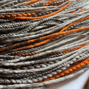 Peut inclure: Un paquet de dreadlocks synth&eacute;tiques dans des tons de gris, brun et orange. Les dreadlocks sont dispos&eacute;es en un motif l&acirc;che et fluide.