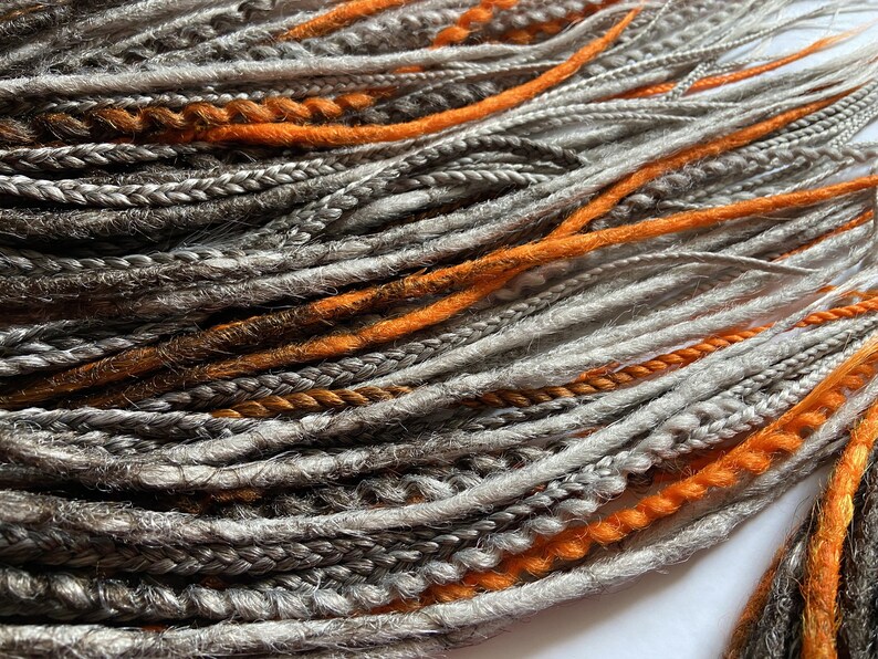 Peut inclure: Un paquet de dreadlocks synth&eacute;tiques dans des tons de gris, brun et orange. Les dreadlocks sont dispos&eacute;es en un motif l&acirc;che et fluide.
