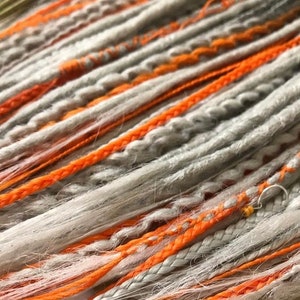 Peut inclure: Dreadlocks synth&eacute;tiques gris et orange. Les dreadlocks sont faites d'un mat&eacute;riau doux et textur&eacute; et ont un aspect naturel, l&eacute;g&egrave;rement d&eacute;sordonn&eacute;.