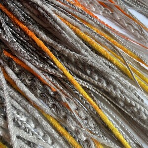 Peut inclure: Une collection de dreadlocks synth&eacute;tiques dans des tons de gris, orange et jaune. Les dreadlocks sont dispos&eacute;es en un motif l&acirc;che et fluide.