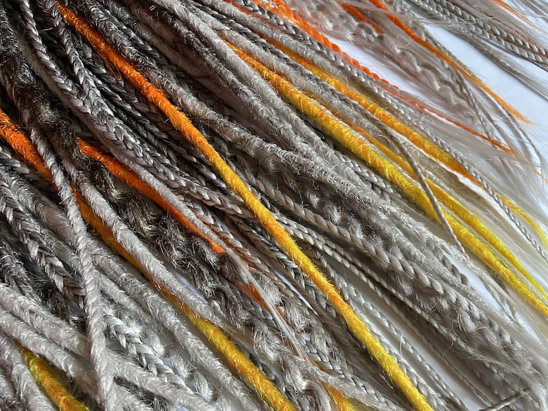 Peut inclure: Une collection de dreadlocks synth&eacute;tiques dans des tons de gris, orange et jaune. Les dreadlocks sont dispos&eacute;es en un motif l&acirc;che et fluide.