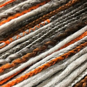 Peut inclure: Gros plan sur une vari&eacute;t&eacute; de dreadlocks synth&eacute;tiques dans des tons de gris, orange et marron. Les dreadlocks sont tress&eacute;es ensemble et ont un aspect textur&eacute; et duveteux.