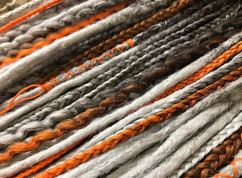 Peut inclure: Gros plan sur une vari&eacute;t&eacute; de dreadlocks synth&eacute;tiques dans des tons de gris, orange et marron. Les dreadlocks sont tress&eacute;es ensemble et ont un aspect textur&eacute; et duveteux.