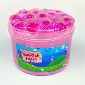 Puede incluir: Un recipiente de plástico transparente lleno de slime rosa brillante y pequeñas cuentas rosas. La etiqueta muestra un fondo azul con colinas verdes y un letrero de madera que dice Jellyfish Fields en rojo, rodeado de burbujas flotantes.
