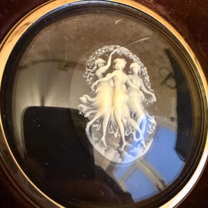 Op de afbeelding: Een witte cameo met drie vrouwen in een cirkel, ingelijst in een goudkleurig metalen frame met een glazen deksel.