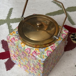 May include: A vintage brass tea caddy with a floral patterned box. The caddy has a round lid and a handle. The box is covered in a colorful floral pattern with pink, yellow, and blue flowers.