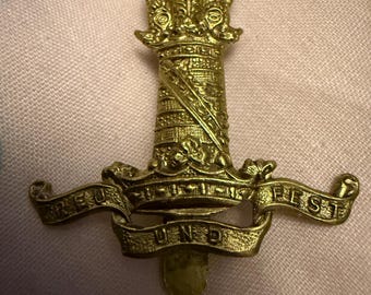 May include: A gold-colored metal badge with a detailed design. The badge features a central column-like structure topped with a decorative element and a banner with text. The overall design is ornate and detailed.