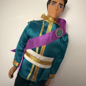 Puede incluir: Mu&ntilde;eco vestido con una chaqueta de sat&eacute;n verde azulado con ribetes dorados y una faja blanca y dorada. Una faja morada est&aacute; drapeada sobre el pecho. El mu&ntilde;eco tiene pelo negro y ojos azules y lleva pantalones negros.