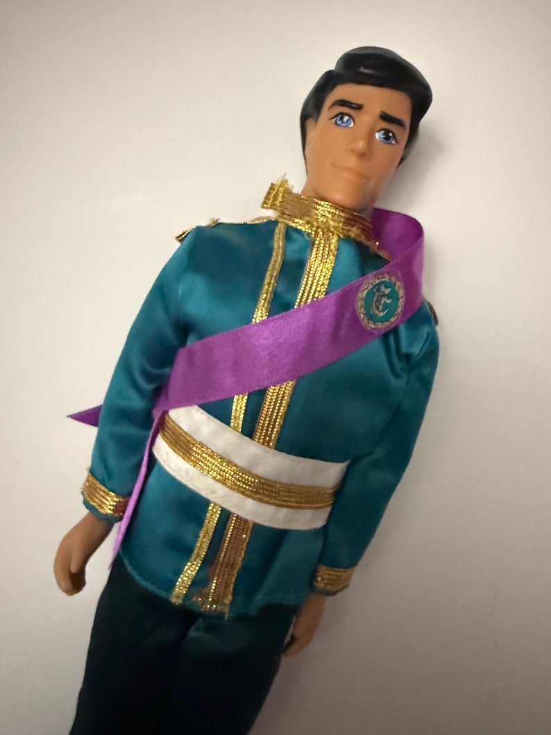 Puede incluir: Mu&ntilde;eco vestido con una chaqueta de sat&eacute;n verde azulado con ribetes dorados y una faja blanca y dorada. Una faja morada est&aacute; drapeada sobre el pecho. El mu&ntilde;eco tiene pelo negro y ojos azules y lleva pantalones negros.