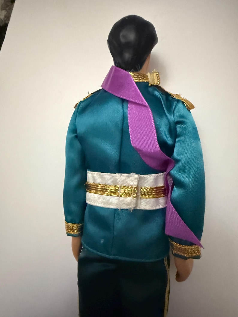 Puede incluir: Mu&ntilde;eca con una chaqueta de sat&eacute;n turquesa con ribetes dorados y charreteras. Una faja morada cruza el pecho. La mu&ntilde;eca tiene pantalones negros y un cintur&oacute;n blanco con ribetes dorados. El pelo de la mu&ntilde;eca es negro.